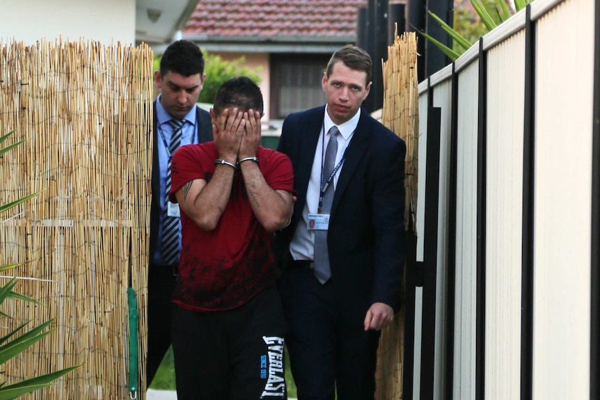 Detectives lead a man in handcuffs through a bamboo screen fence.