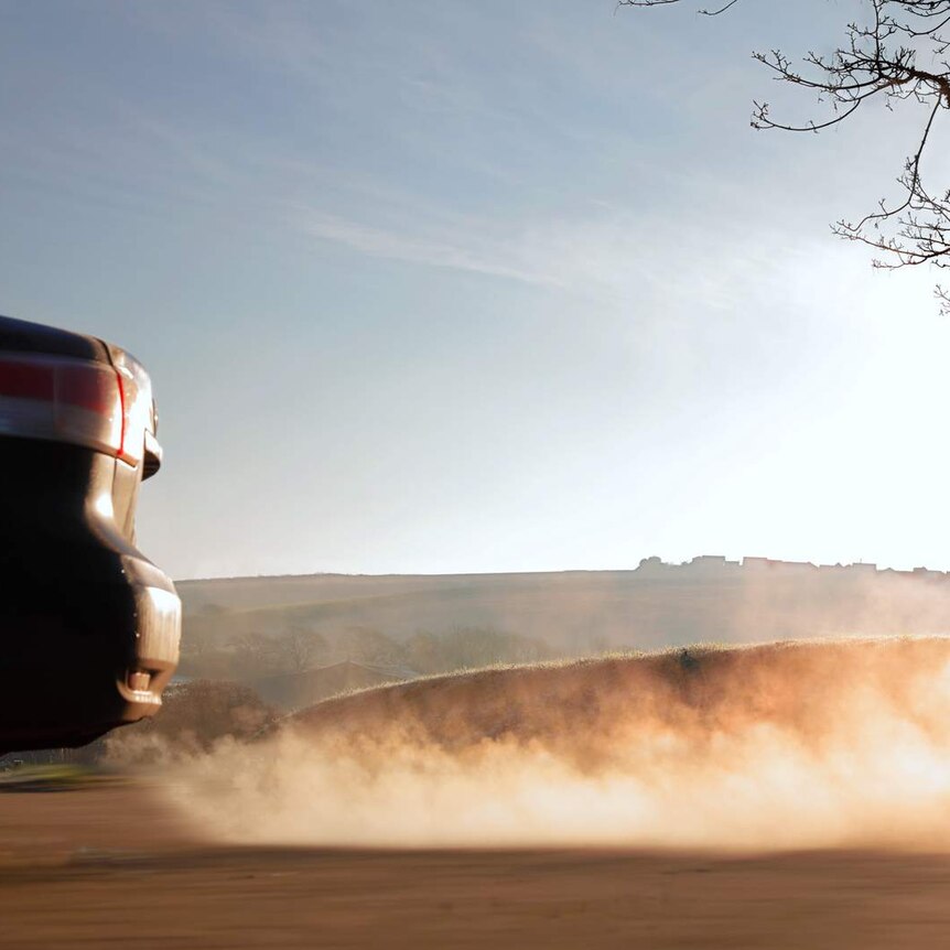 A car drives along a country road and exhaust fumes visibly trail behind, captured in the sunlight in front of a hill.