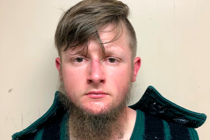 This booking photo shows a young man with a long beard and long fringe looking at the camera.