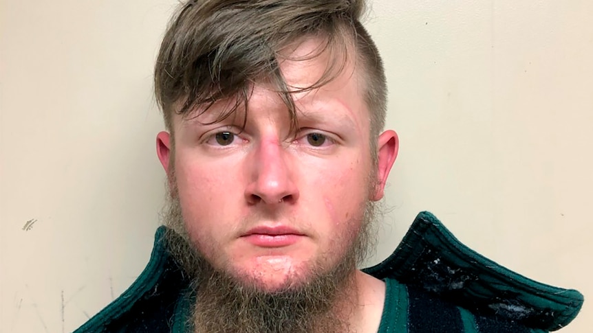 This booking photo shows a young man with a long beard and long fringe looking at the camera.