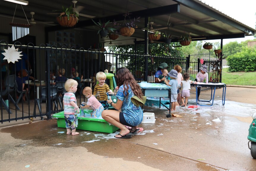 Kids at a daycare play outside in water. There is teachers talking to them and foam.