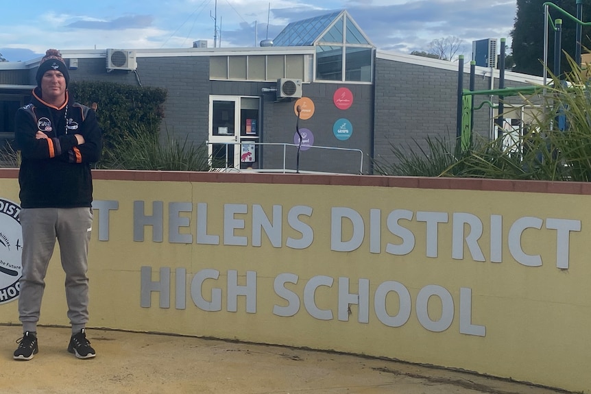 A man wearing a beanie stands in front of the St Helens District High School 