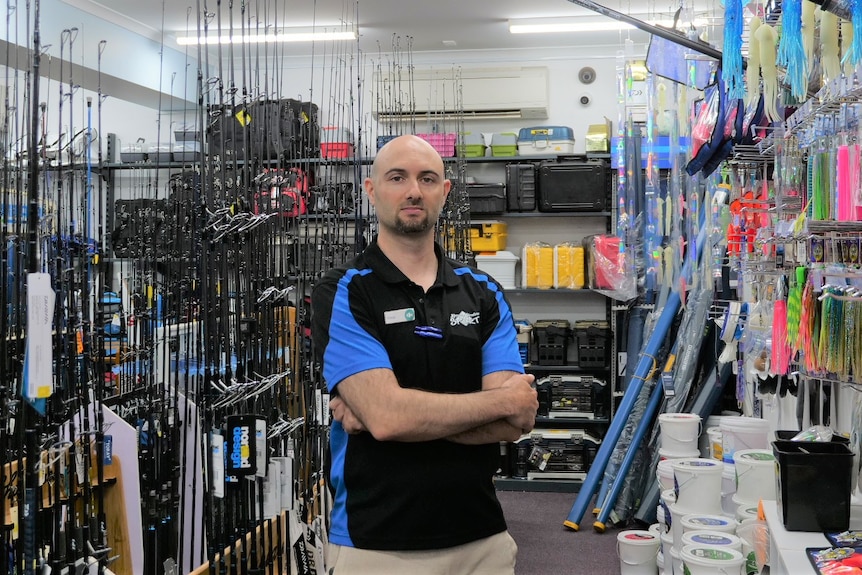 A man stands in between aisles with his arms crossed in a fishing store. Fishing rods are