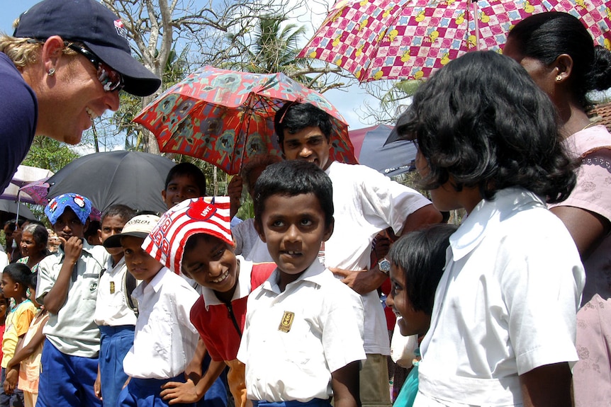 Shane Warne leans down to speak with Sri Lankan children. Behind them, adults hold bright umbrellas to block out sun