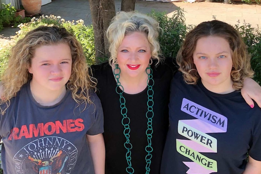 A woman with blond hair with her arms around two teenage girls while standing outside.