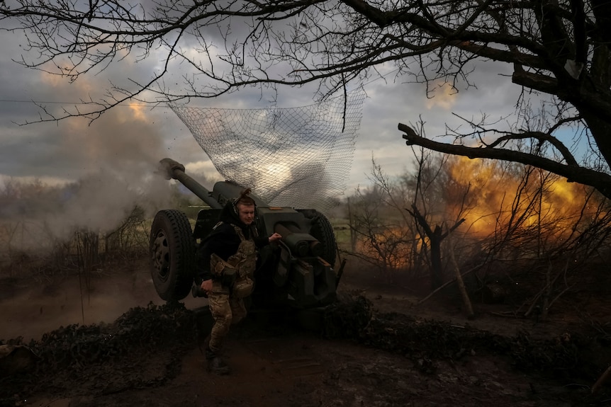 Ukrainian service members fire a howitzer D30 at a front line as flames can be seen in the bushes nearby. 
