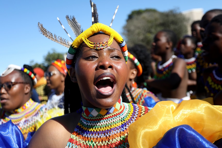 A woman is dressed in a traditional colorful Zulu headdress and necklace.  Others are dressed in bright colors behind her. 