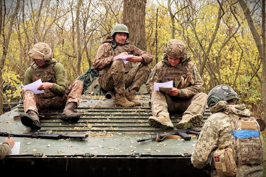 Three men in military uniforms sitting on a tank, writing on paper, in a forrest.