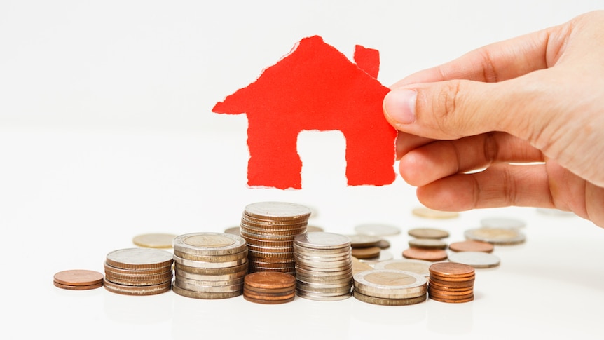 A hand holds a cutout of a red house above a pile of coins.