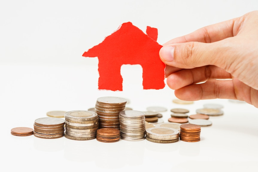 A hand holds a cutout of a red house above a pile of coins.