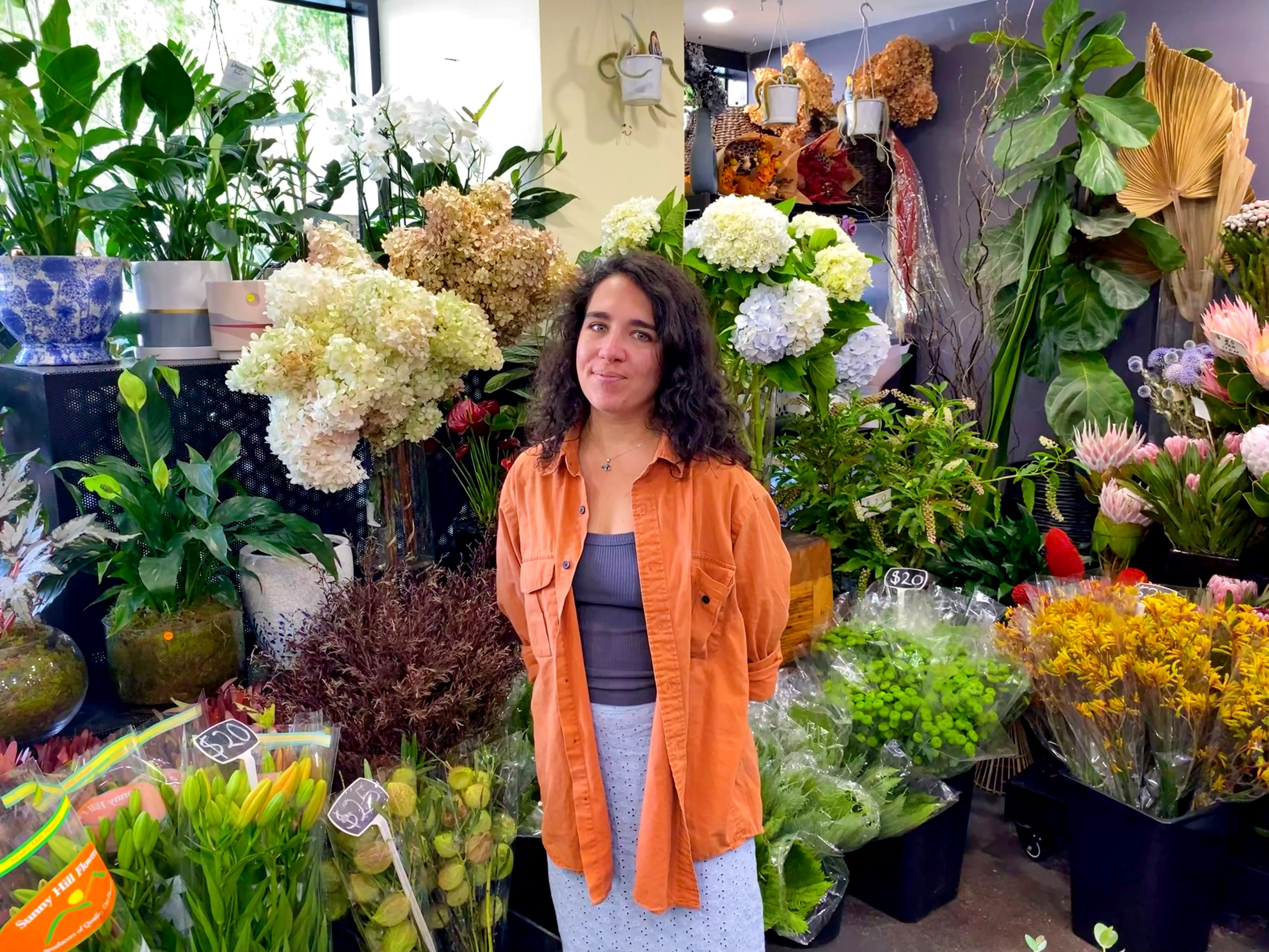 Uma mulher de cabelos escuros está entre buquês de flores em uma loja.