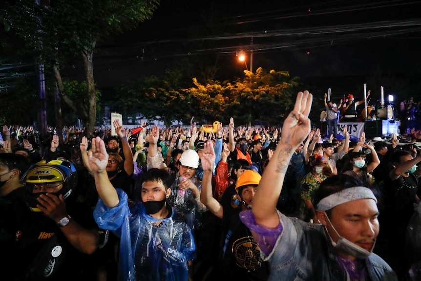 A group of people wearing raincoats and masks sit on the ground holding up three fingers in the air.