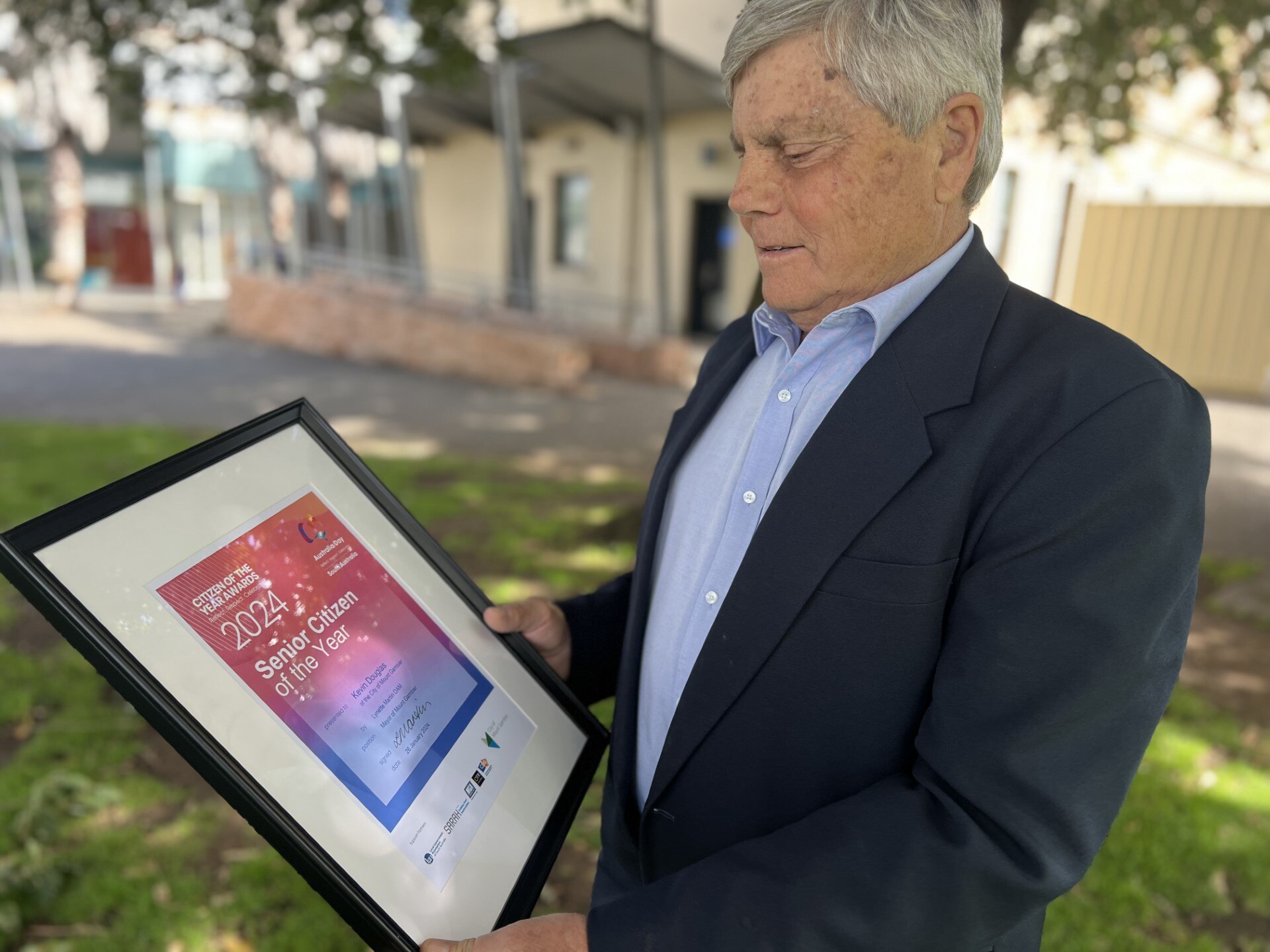 A grey haired man in blue blazer holds framed Australia Day award