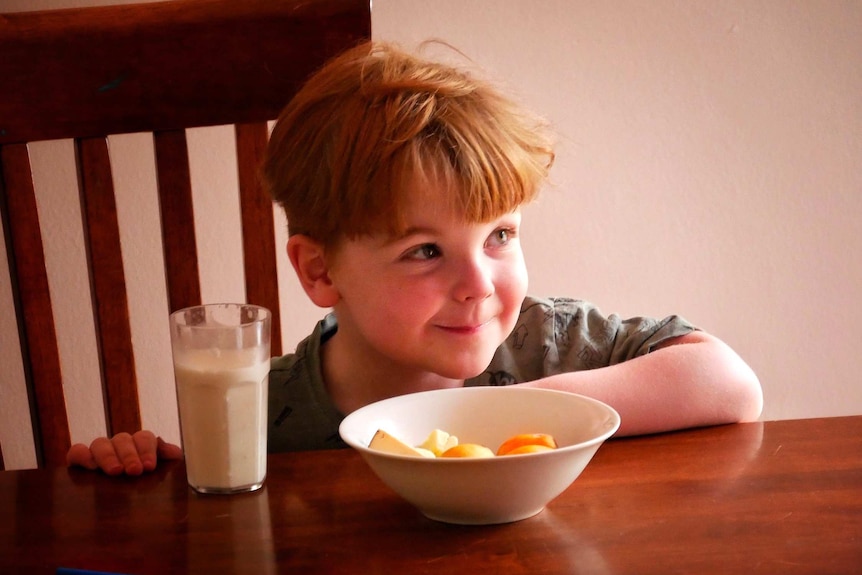 A young boy smiles as he sits at a table with a bowl of fruit and glass of milk in front of him.