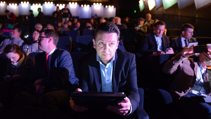 Nick Xenophon sits in a chair in a cinema holding a tablet device.