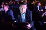 Nick Xenophon sits in a chair in a cinema holding a tablet device.