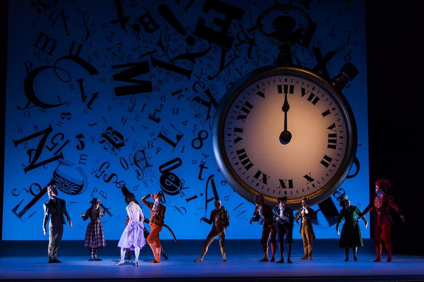 Eleven dancers in colourful costumes perform on stage in front of a giant clock.