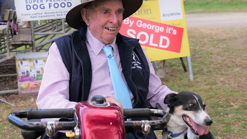 An old man in a hat, beaming, sits in a motor scooter next to a black and white dog.
