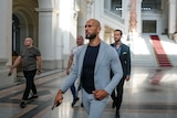 A bald man wearing a grey suit and tshirt walking inside a big marble room with three people behind him