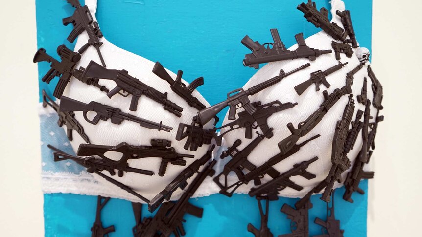 A mixed media artwork featuring a bra covered in tiny guns.