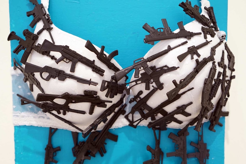 A mixed media artwork featuring a bra covered in tiny guns.