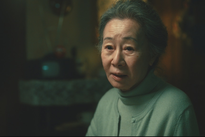An elderly  Korean woman in a pale green turtle neck and cardigan looks pensively at the camera. 