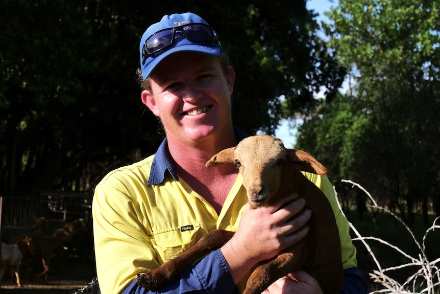 A man wearing high-vis clothing and a cap in a paddock, holding a small goat.