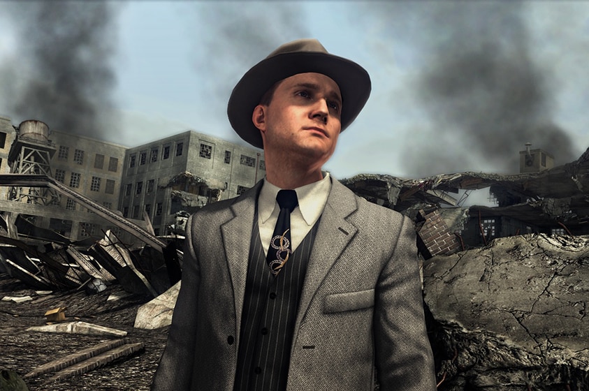 In a scene from a video game a man in American 1940s hat and suit stands among building ruins as black smoke rises to the sky.