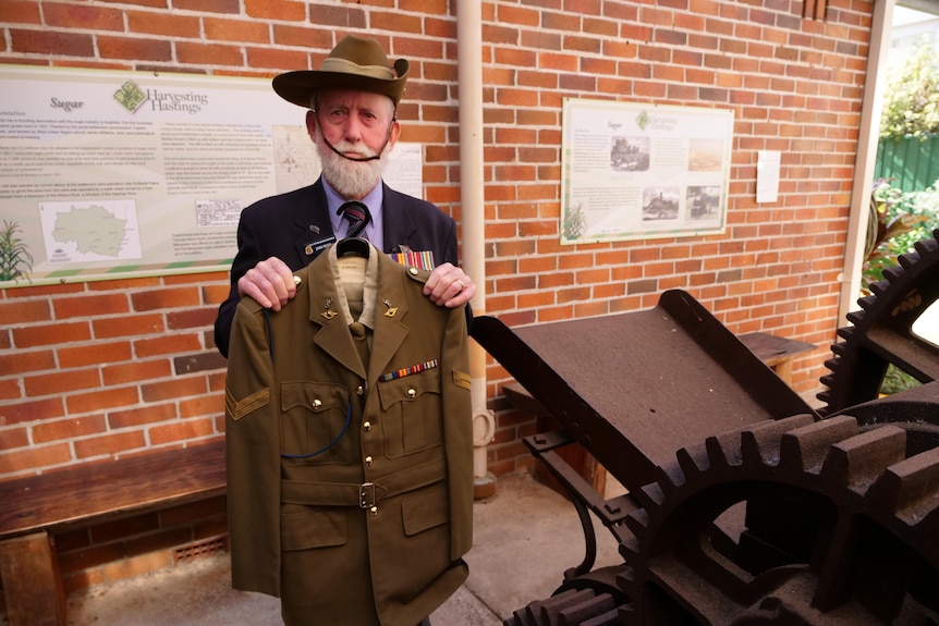 A man holding up his army uniform in front of a brick wall. 