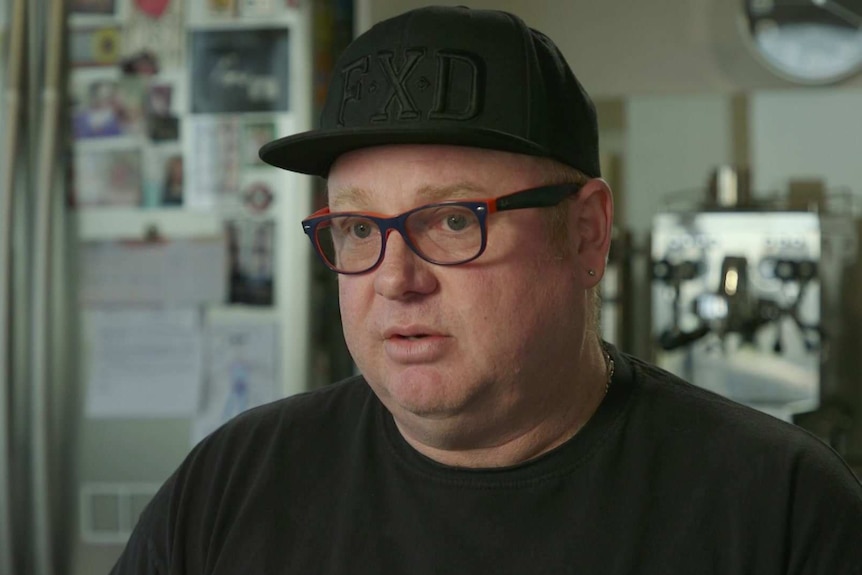 Head shot of Hugo Armstrong wearing a black baseball-style cap with FXD on the front. Blurry kitchen scene in the background