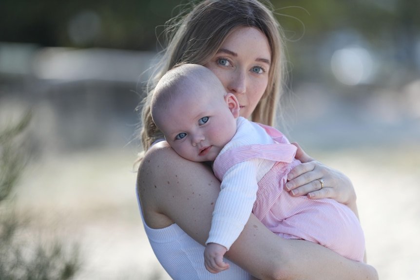 Sarah stands in focus on a beach holding her baby