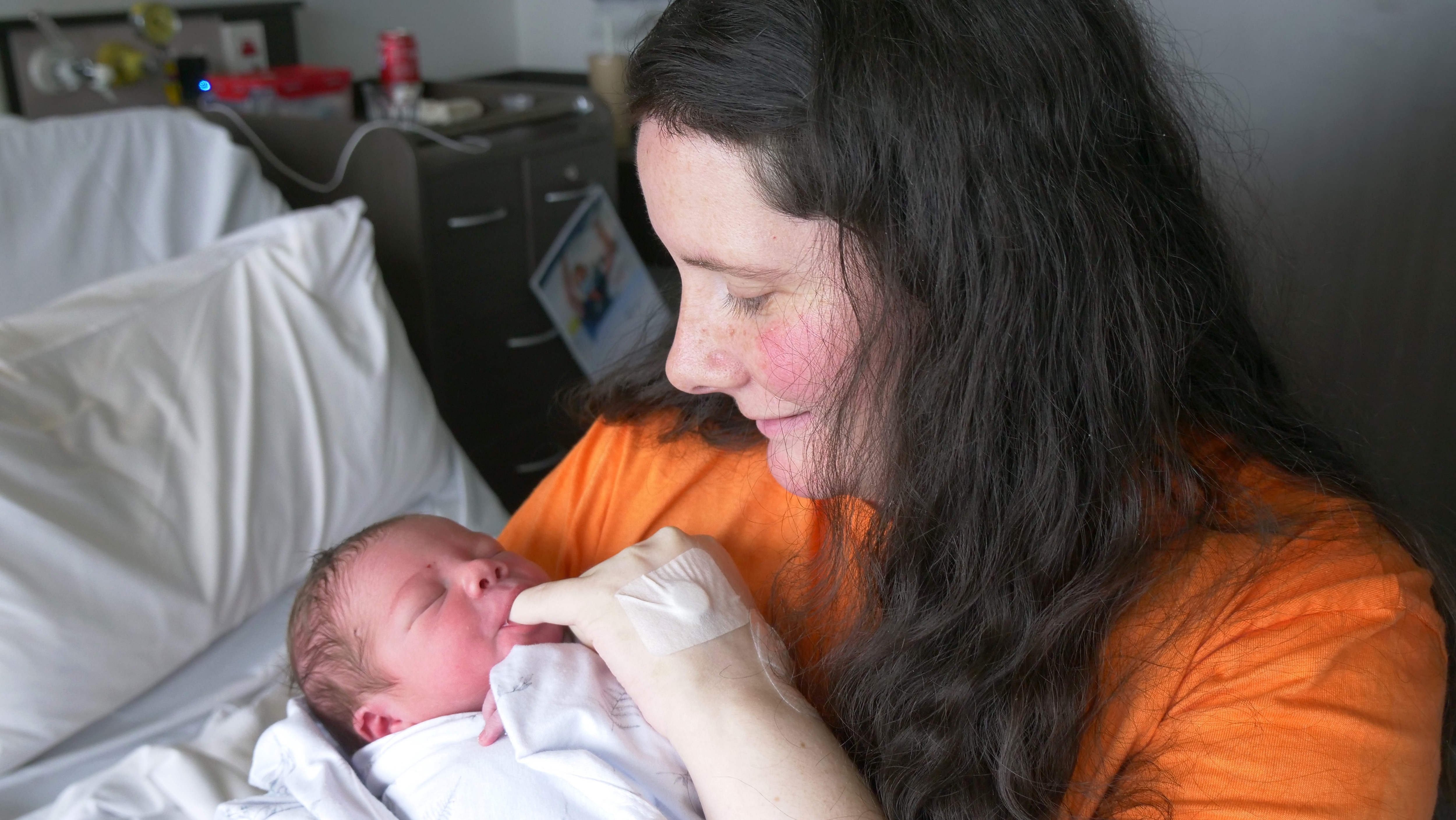 A woman in an orange shirt and a baby in a wrap sit on a hospital bed.