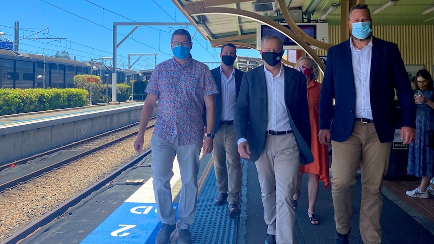 A group of men and women walk along a railway platform, wearing masks.