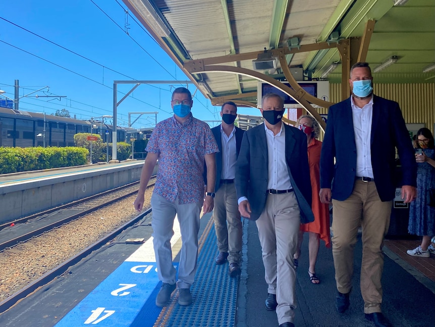 A group of men and women walk along a railway platform, wearing masks.