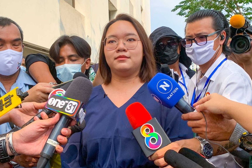 A young woman stands surrounded by journalists holding microphones under her face