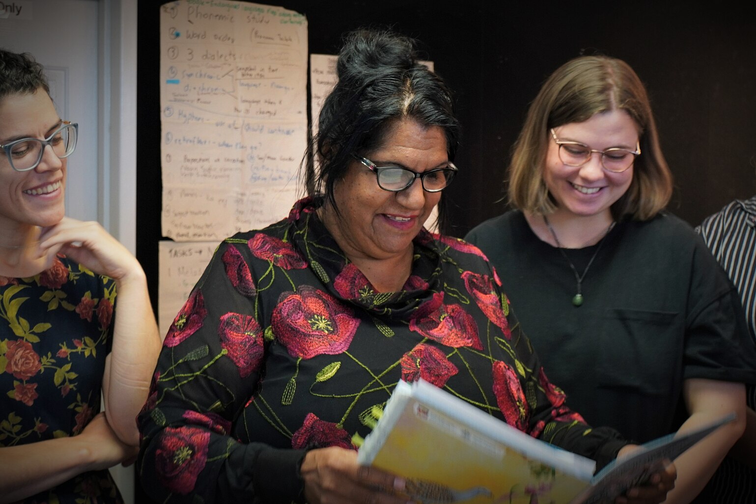 A woman with black hair and glasses wearing a floral shirt while two other women look at a document over her shoulder