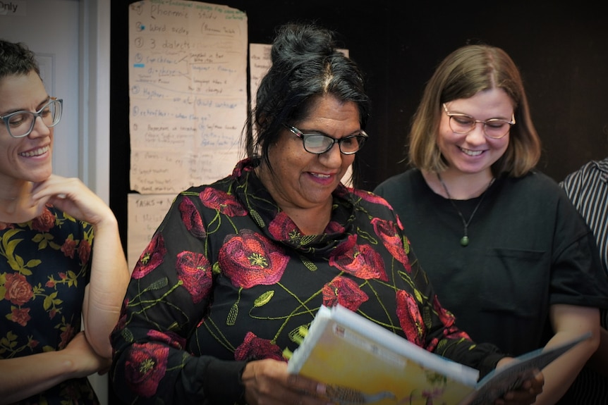 A woman with black hair and glasses wearing a floral shirt while two other women look at a document over her shoulder