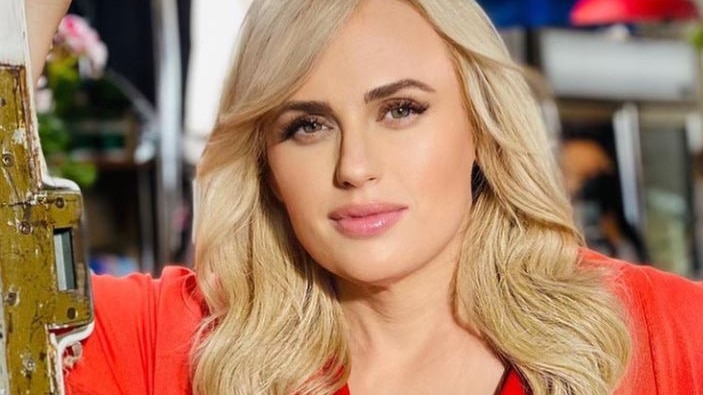 Rebel Wilson stands in front of a door and looks to the camera with a soft expression. Her blonde hair's down, she wears red