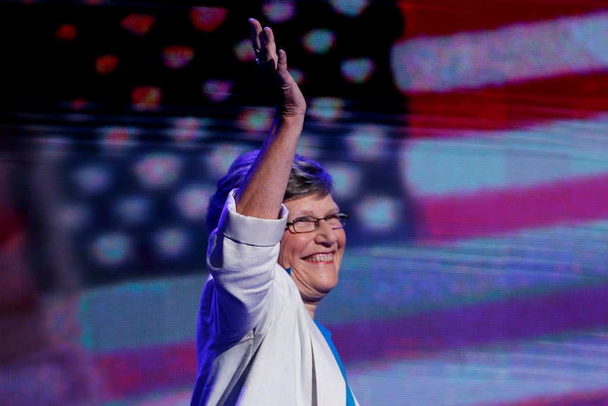 sister simone campbell waves her arm with a big smile on her face. an american flag banner can be seen behind her