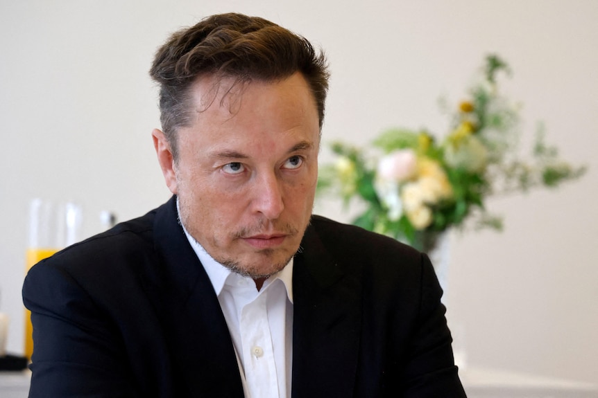 A portrait photo of Elon Musk. He is a white man with short black hair and brown eyes, wearing a black suit. 