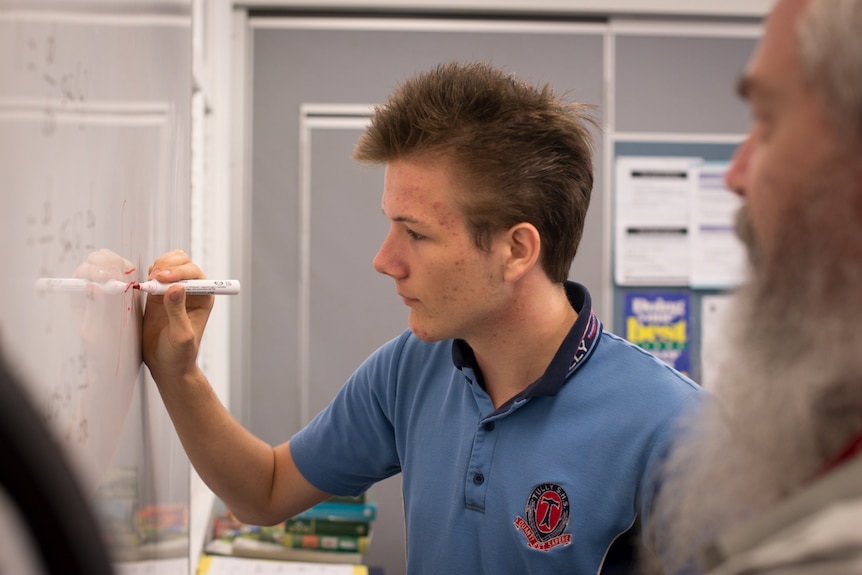 A high school student works on a whiteboard while a bearded teacher looks on