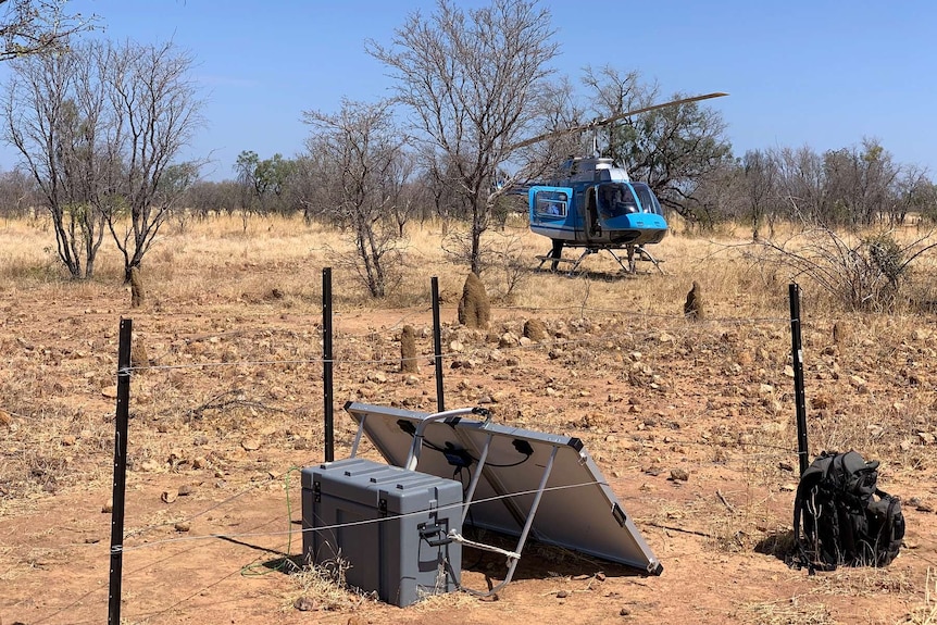A grey box and solar panel is seen at the base of a fence line in the Beetaloo Basin. A blue helicopter is parked behind.