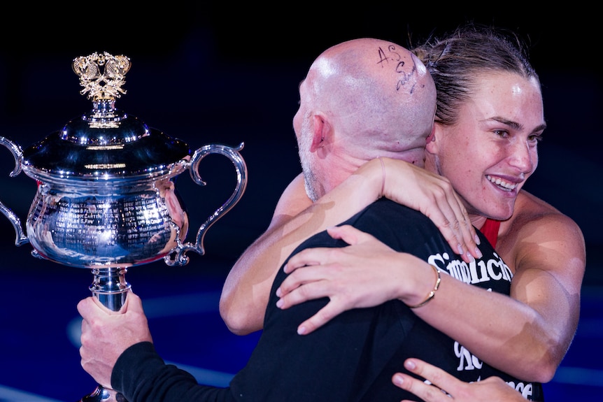 Aryna Sabalenka hugs trainer Jason Stacy after the Australian Open final. He is holding the trophy.