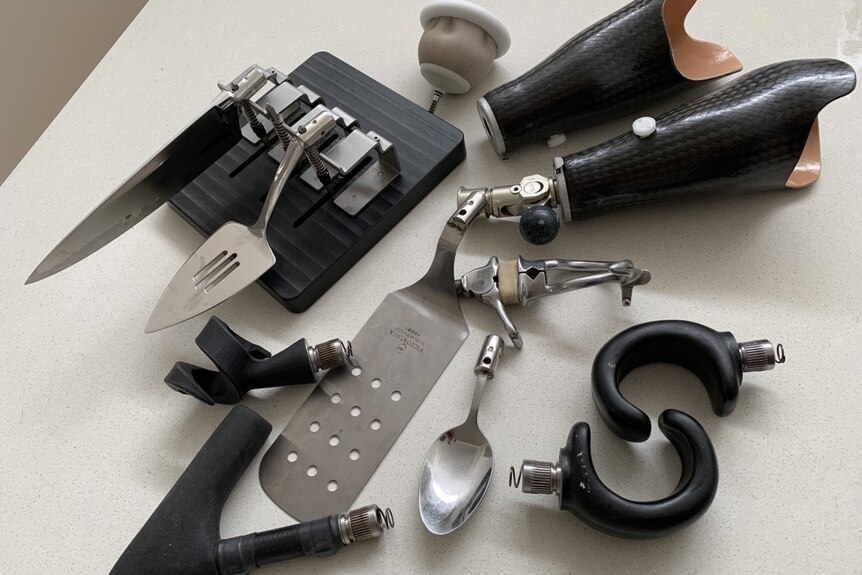 A selection of modified metal kitchen utensils, cutlery and hooks sit on a kitchen bench next to prosthetic limbs