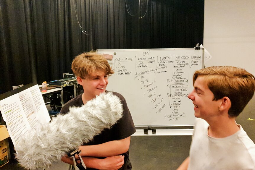 Two blonde-haired teenage boys stand laughing in a room, with a boom mic between them, as they speak into it.