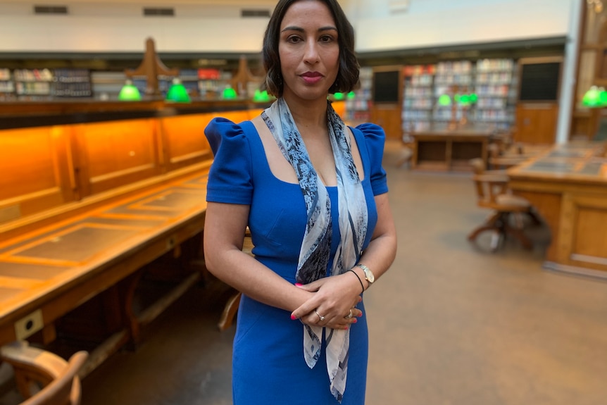 Dr Emma Fulu wearing a blue dress, standing in a library with her hands folded. 