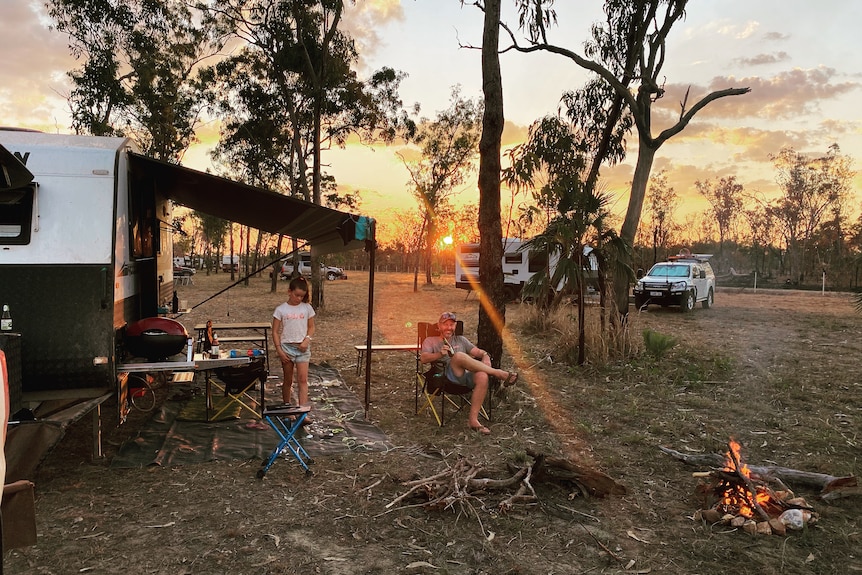 Orange low sun and trees behind a man in camping chair, smiling, holding a beer, and young girl and caravan to his side.