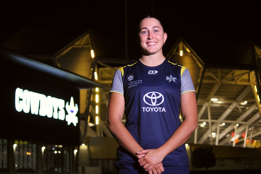 A woman wearing a football jersey stands in front of an illuminated stadium and Cowboys sign at night 