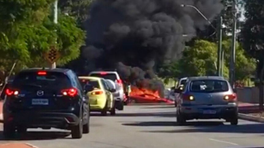Thick black smoke rises into the sky from a burning Ferrari on a road with other cars stopped in the foreground.