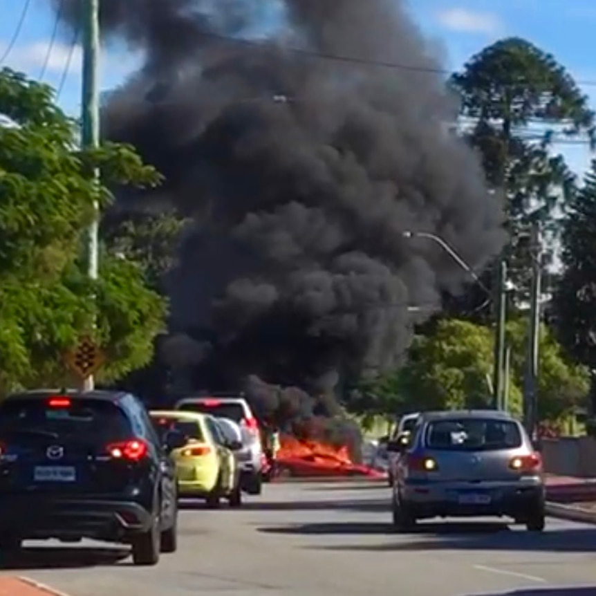Thick black smoke rises into the sky from a burning Ferrari on a road with other cars stopped in the foreground.